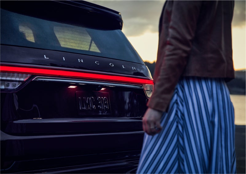 A person is shown near the rear of a 2023 Lincoln Aviator® SUV as the Lincoln Embrace illuminates the rear lights | Buss Lincoln in McHenry IL