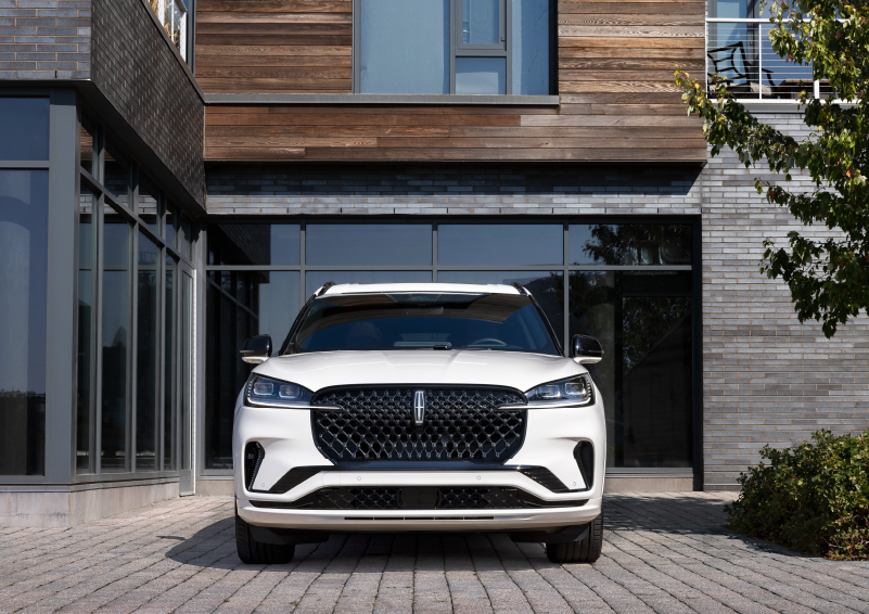 A white 2025 Lincoln Aviator® SUV is shown parked outside a modern building with gray brick and wood accents. | Buss Lincoln in McHenry IL