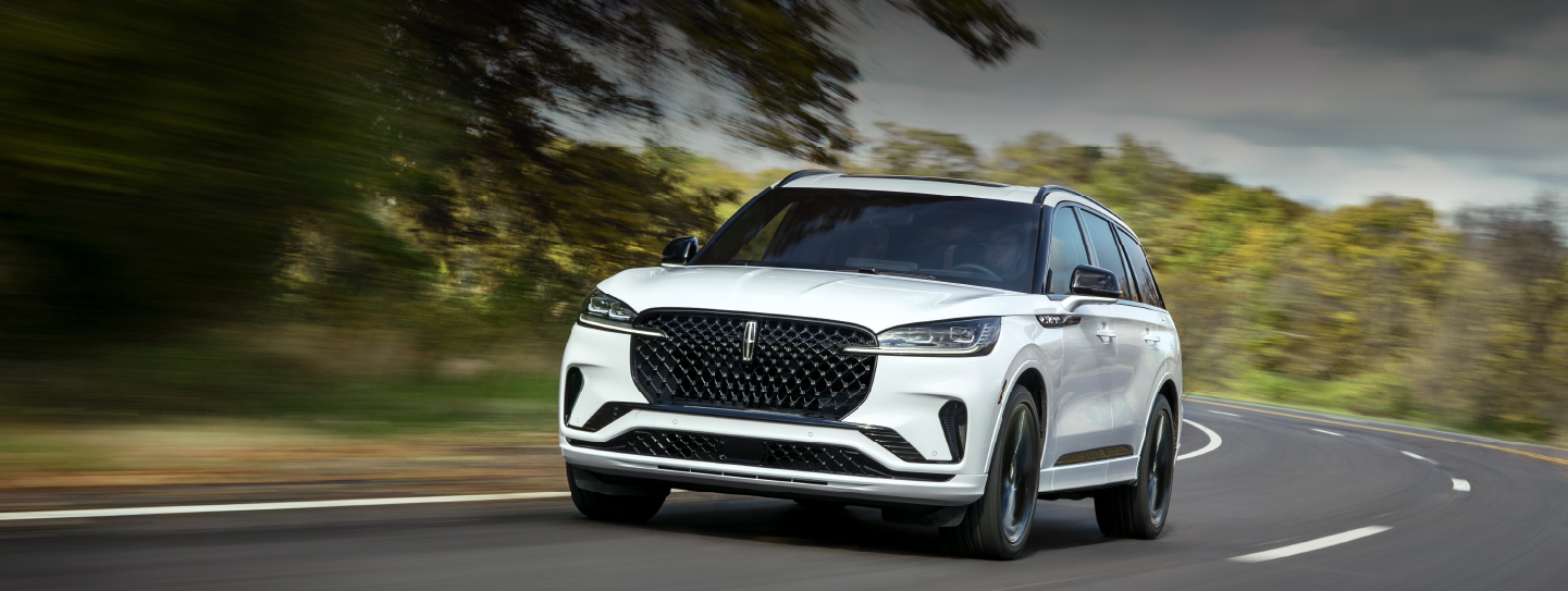 A white 2025 Lincoln Aviator® SUV is being driven down a road lined with trees. | Buss Lincoln in McHenry IL