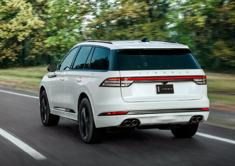 A white 2026 Lincoln Aviator® SUV is being driven away down a road lined with colorful trees. | Buss Lincoln in McHenry IL